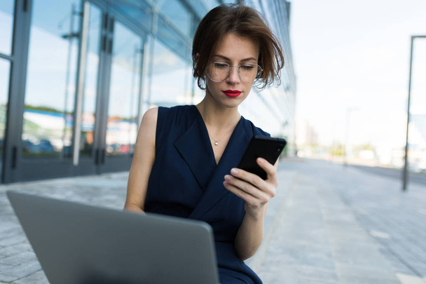 femme d'affaires travaillant avec ordinateur portable et téléphone à l'extérieur près du bâtiment moderne
  - Photo, image