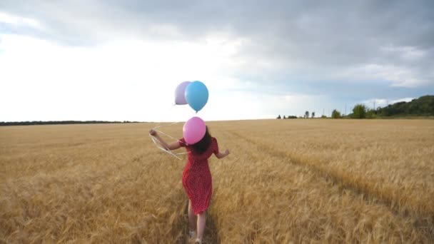 Szczęśliwa dziewczyna w czerwonej sukience biegnie przez złote pole pszenicy z balonami w ręku w pochmurny dzień. Młoda kobieta z brązowymi włosami bawiąca się podczas joggingu wśród plantacji jęczmienia. Koncepcja wolności - Materiał filmowy, wideo