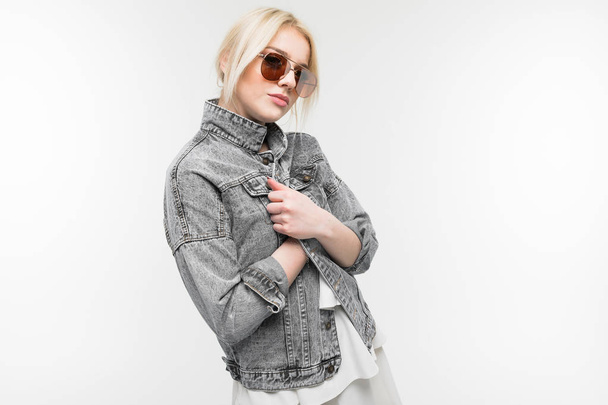 portrait of young charming joyful blonde girl in jeans jacket possing and gesticulating in round sunglasses on background - Zdjęcie, obraz