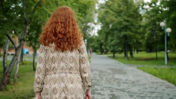 Back view of pretty redhead girl walking in park alone turning to camera smiling - Filmagem, Vídeo