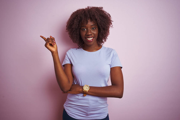 Молодая африканская женщина в футболке, стоящая на изолированном розовом фоне с большой улыбкой на лице, указывая рукой и пальцем в сторону, глядя в камеру
. - Фото, изображение