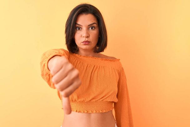 Νεαρή όμορφη γυναίκα φορώντας casual t-shirt στέκεται πάνω από απομονωμένο πορτοκαλί φόντο φαίνεται δυστυχισμένη και θυμωμένη δείχνει απόρριψη και αρνητική με τους αντίχειρες προς τα κάτω χειρονομία. Κακή έκφραση. - Φωτογραφία, εικόνα