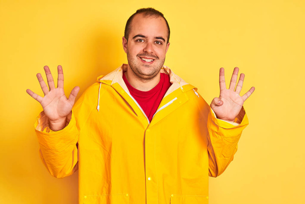 Jeune homme portant un imperméable debout sur fond jaune isolé montrant et pointant vers le haut avec les doigts numéro neuf tout en souriant confiant et heureux
. - Photo, image