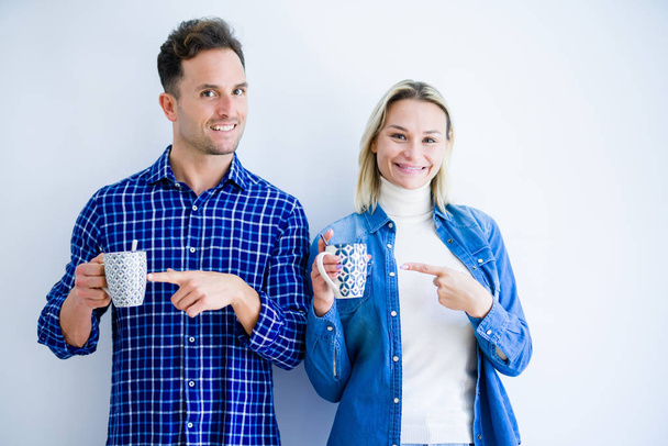 Nuori kaunis pari juominen kuppi kahvia seisoo yli eristetty valkoinen tausta erittäin onnellinen osoittaa käsin ja sormella
 - Valokuva, kuva
