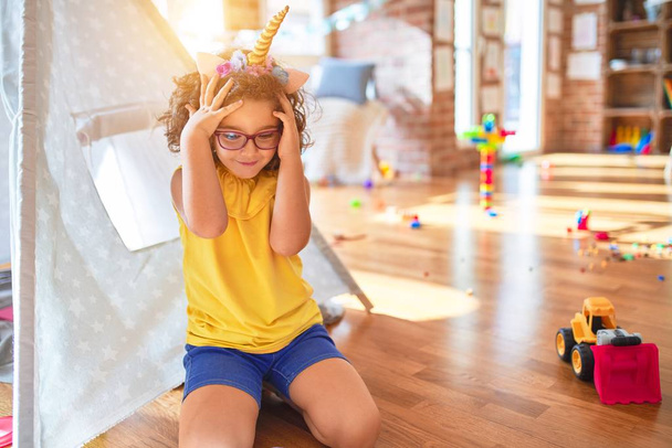 Beautiful toddler wearing glasses and unicorn diadem sitting on the floor at kindergarten suffering from headache desperate and stressed because pain and migraine. Hands on head. - Foto, immagini