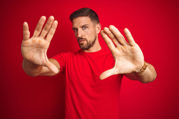 Kırmızı izole arka plan üzerine günlük tişört giyen genç yakışıklı adam el ve parmakları kullanarak çerçeve yapıyor, kamera perspektifi kullanıyor. - Fotoğraf, Görsel