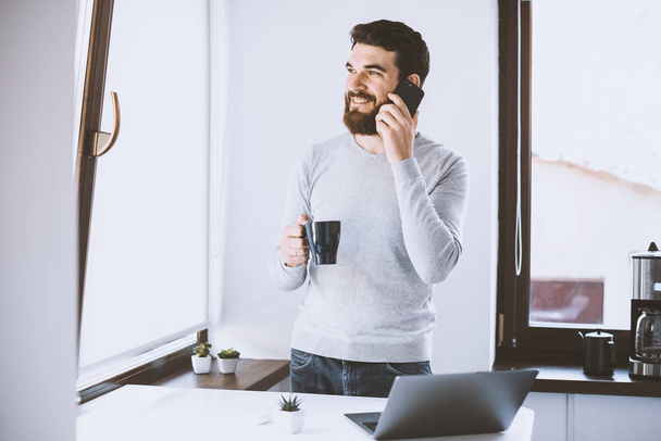 Foto van vrolijke jonge casual man praten op telefoon en het drinken van zijn ochtend koffie - Foto, afbeelding