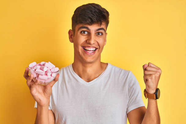 Young indian man holding bowl with marshmallows over isolated yellow background very happy and excited, winner expression celebrating victory screaming with big smile and raised hands - Photo, Image
