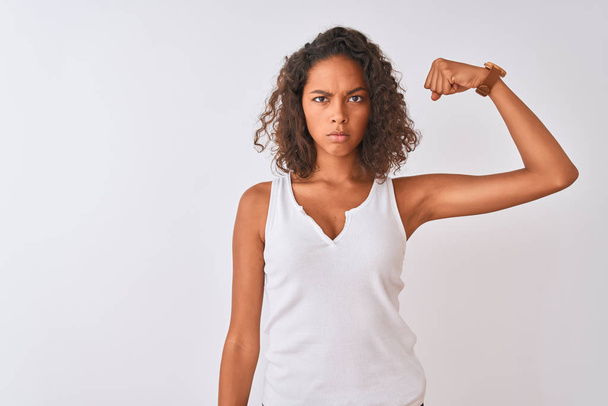 junge Brasilianerin in lässigem T-Shirt vor isoliertem weißem Hintergrund starke Person, die Armmuskeln zeigt, selbstbewusst und stolz auf ihre Macht - Foto, Bild