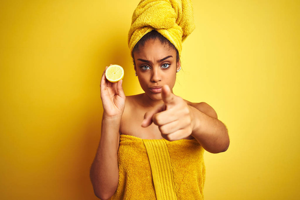 Afro γυναίκα φορώντας πετσέτα μετά το ντους κρατώντας φέτα λεμόνι πάνω από απομονωμένο κίτρινο φόντο δείχνοντας με το δάχτυλο στην κάμερα και σε σας, σημάδι στο χέρι, θετική και σίγουρη χειρονομία από το μπροστινό μέρος - Φωτογραφία, εικόνα