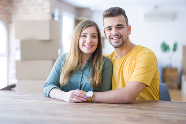 Молода красива пара сидить на столі вдома, обіймаючись любов'ю дуже щаслива за переїзд в новий будинок з картонними коробками за ними
 - Фото, зображення