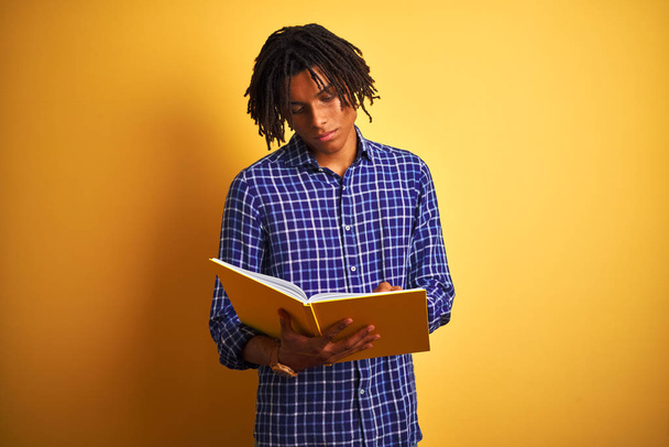 アフリカ系アメリカ人学生男とともにdrellocks読書本上の隔離された黄色の背景とともに自信を持って式上のスマート顔考え深刻な - 写真・画像