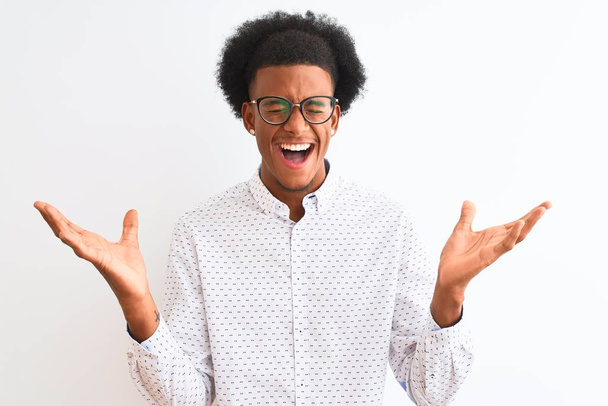 Νεαρός Αφροαμερικάνος που φοράει κομψό πουκάμισο και γυαλιά πάνω από απομονωμένο λευκό φόντο γιορτάζει τρελός και τρελός για την επιτυχία με τα χέρια ψηλά και κλειστά μάτια ουρλιάζοντας ενθουσιασμένος. Έννοια νικητή - Φωτογραφία, εικόνα