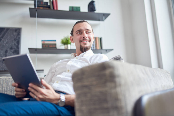 Photo of happy brunet looking to side with tablet in his hands sitting on sofa in room . - Fotoğraf, Görsel
