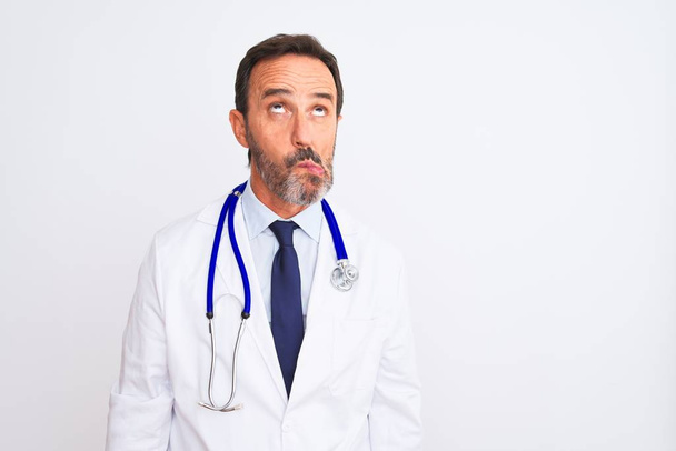 Lekarz w średnim wieku, noszący płaszcz i stetoskop stojący nad odizolowanym białym tłem, robiący rybią twarz ustami, szalony i komiczny gest. Śmieszne wyrażenie. - Zdjęcie, obraz