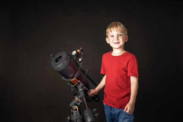 赤いフットボールの小さな男の子が大きな望遠鏡の隣に立っている. - 写真・画像