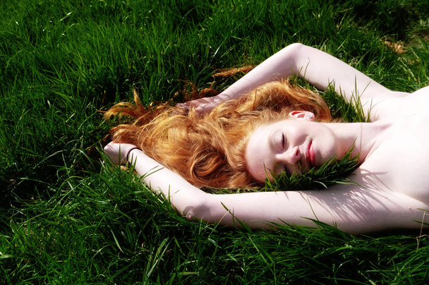 若いセクシーな赤髪の女性の美しい肖像画、彼女の腕を上げて、緑の芝生の上に夏の太陽の下で横たわって、赤い髪は頭の周りに自由にドレープ. - 写真・画像