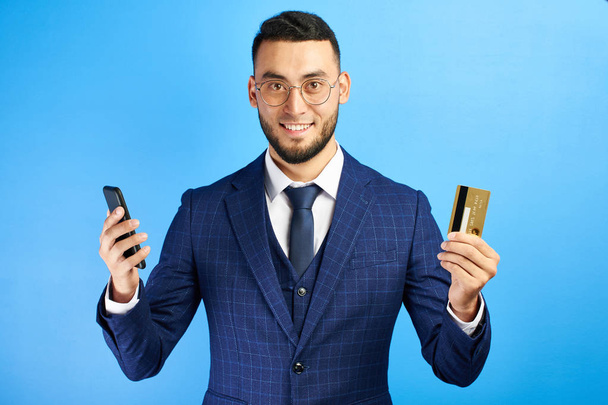 Asian Kazakh business bank representative man in suit and glasses smiles joyfully and holds mobile phone and credit card in hands on blue background isolated - Photo, Image