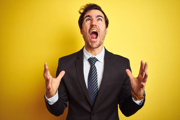 Young handsome businessman wearing suit and tie standing over isolated yellow background crazy and mad shouting and yelling with aggressive expression and arms raised. Frustration concept. - Photo, image