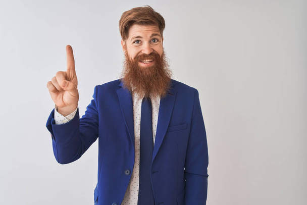 Young redhead irish businessman wearing suit standing over isolated white background showing and pointing up with finger number one while smiling confident and happy. - Photo, image