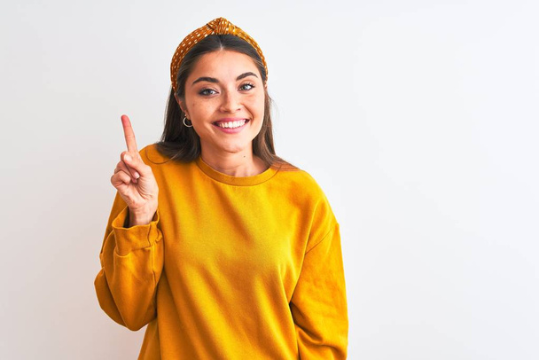 Mujer hermosa joven con suéter amarillo y diadema sobre fondo blanco aislado que muestra y señala hacia arriba con el dedo número uno mientras sonríe confiado y feliz
. - Foto, imagen