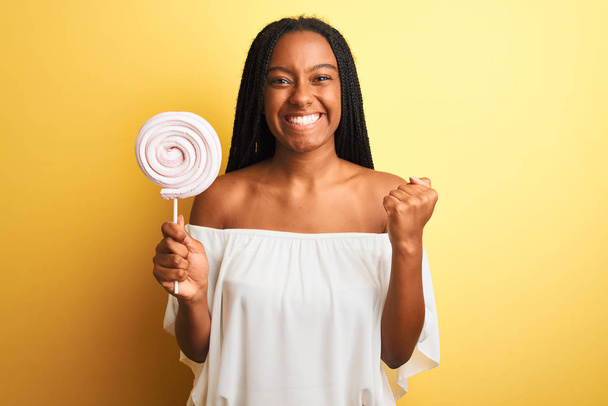 Jovem afro-americana comendo doces em pé sobre fundo amarelo isolado gritando orgulhoso e celebrando vitória e sucesso muito animado, animando emoção
 - Foto, Imagem