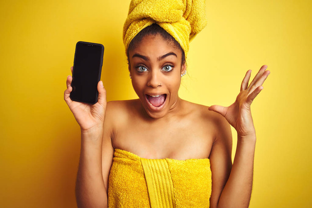 Afro femme portant serviette après douche montrant smatrphone sur fond jaune isolé très heureux et excité, expression gagnante célébrant la victoire en criant avec un grand sourire et les mains levées
 - Photo, image