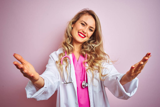 Young beautiful doctor woman using stethoscope over pink isolated background looking at the camera smiling with open arms for hug. Cheerful expression embracing happiness. - Foto, Imagen