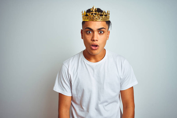 Young brazilian man wearing king crown standing over isolated white background afraid and shocked with surprise expression, fear and excited face. - Фото, зображення
