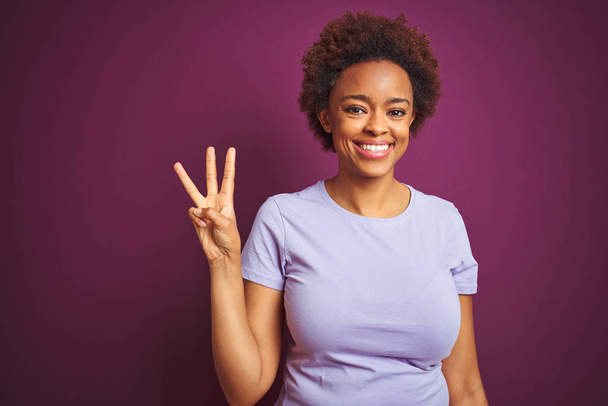 Νεαρή όμορφη Αφροαμερικανή γυναίκα με αφρο μαλλιά πάνω από απομονωμένο πορφυρό φόντο που δείχνει και δείχνει προς τα πάνω με τα δάχτυλα νούμερο τρία, ενώ χαμογελά αυτοπεποίθηση και χαρούμενος. - Φωτογραφία, εικόνα