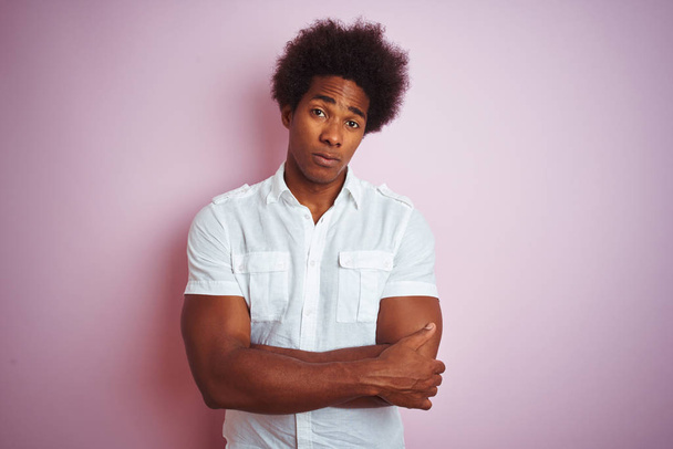 Молодий американський чоловік з волоссям афроамериканського походження, одягнений в білу сорочку, стоїть над ізольованим рожевим фоном скептично і нервово, не схвалюючи виразу на обличчі перехрещеними руками. Негативна людина. - Фото, зображення