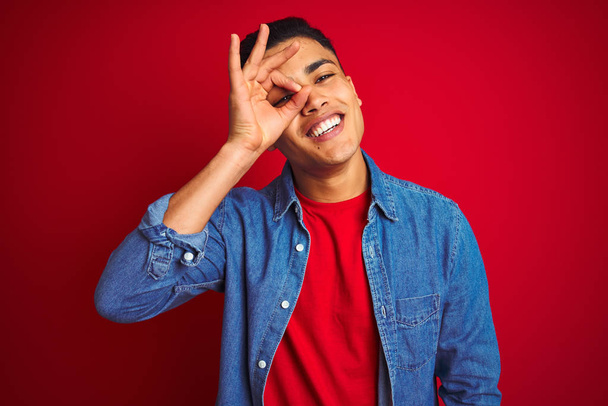 Jonge Braziliaanse man draagt denim shirt staan over geïsoleerde rode achtergrond doen ok gebaar met de hand glimlachen, oog kijken door vingers met gelukkig gezicht. - Foto, afbeelding