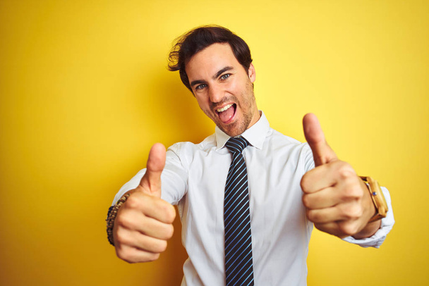 Der junge, gut aussehende Geschäftsmann trägt elegantes Hemd und Krawatte über gelbem Hintergrund und zustimmend positive Gesten mit der Hand, hebt lächelnd die Daumen und freut sich über den Erfolg. Siegergeste. - Foto, Bild