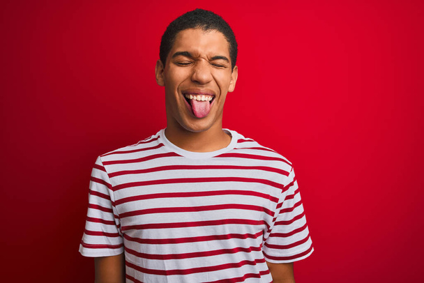 Young handsome arab man wearing striped t-shirt over isolated red background sticking tongue out happy with funny expression. Emotion concept. - Foto, afbeelding