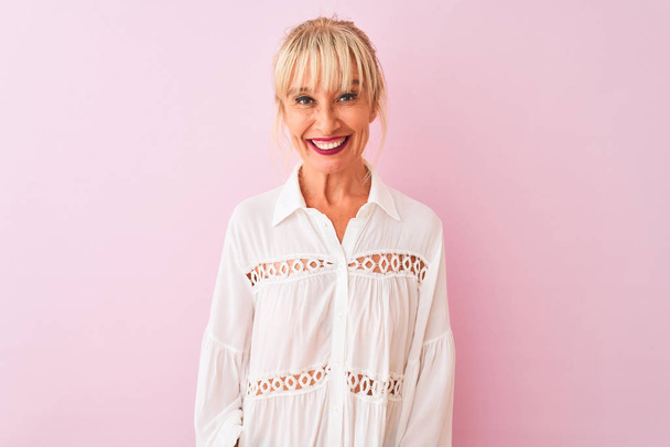 Жінка середнього віку в білій повсякденній сорочці стоїть над ізольованим рожевим тлом з щасливою і прохолодною посмішкою на обличчі. Щаслива людина
. - Фото, зображення