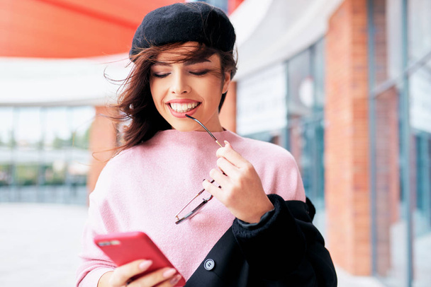 Close-up image of smiling happy woman standing outdoors texting message on mobile phone. Young female wears pink sweater, black beret with blowing hair, browsing on her cell phone on the city street - Foto, Imagen