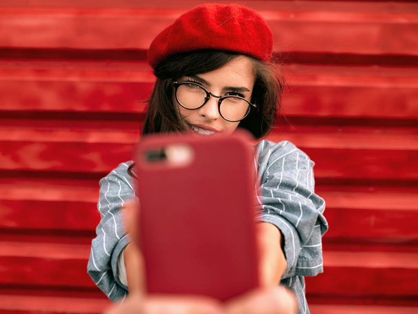 Close-up portrait of brunette young woman smiling broadly, taking self portrait, wearing blue shirt, red beret hat and glasses, posing on red background wall. Student female making selfie on cellphone - Photo, image
