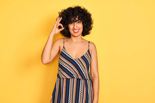 Arab woman with curly hair wearing striped colorful dress over isolated yellow background smiling positive doing ok sign with hand and fingers. Successful expression. - Valokuva, kuva