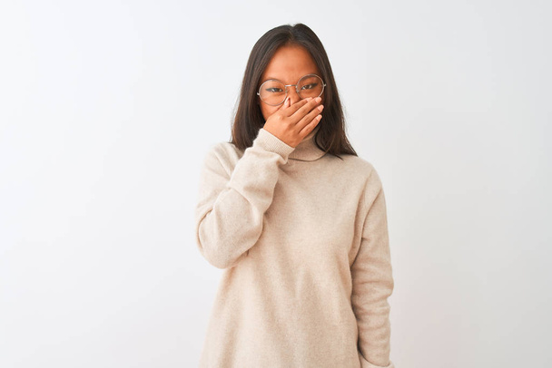 Jonge Chinese vrouw draagt een trui met coltrui en een bril over een geïsoleerde witte achtergrond en ruikt iets stinkends en walgelijks, een ondraaglijke geur, houdt adem in met vingers op haar neus. Slecht geurconcept. - Foto, afbeelding