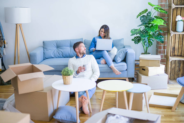 Young beautiful couple sitting on the sofa drinking cup of coffee using laptop at new home around cardboard boxes - Zdjęcie, obraz
