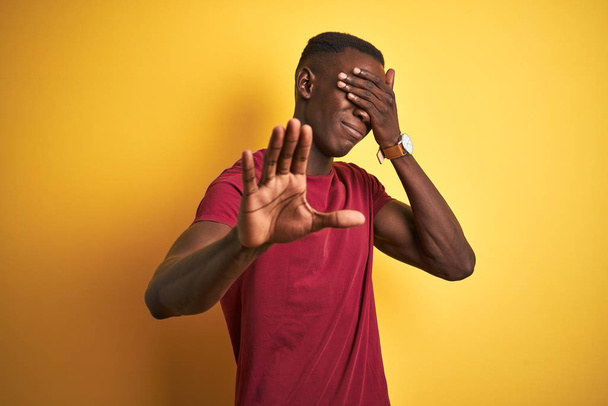 Młody, afrykański Amerykanin w czerwonej koszulce, stojący nad odizolowanym żółtym tłem pokrywającym dłońmi oczy i wykonujący gest zatrzymania ze smutkiem i strachem. Zawstydzona i negatywna koncepcja. - Zdjęcie, obraz