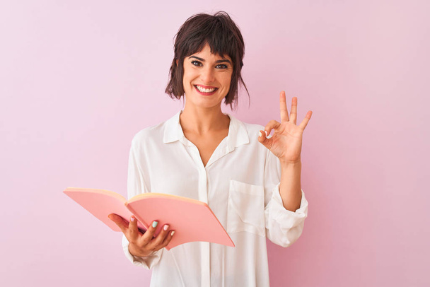 Молодая красивая учительница женщина читает книгу стоя на изолированном розовом фоне делает хорошо знак с пальцами, отличный символ
 - Фото, изображение
