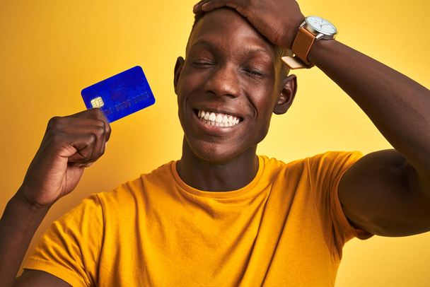 Afrikai amerikai ügyfél férfi kezében hitelkártya felett áll elszigetelt sárga háttér stresszes kézzel a fejét, sokkolt a szégyen és meglepetés arc, dühös és frusztrált. Félelem és tévedés. - Fotó, kép