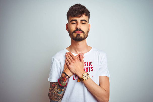 Молодой человек с татуировкой в поясной футболке стоял на изолированном белом фоне улыбаясь руками на груди с закрытыми глазами и благодарным жестом на лице. Концепция здоровья
. - Фото, изображение