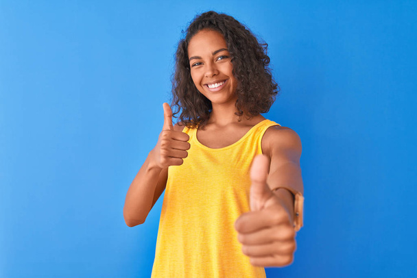 Jonge Braziliaanse vrouw met geel t-shirt over geïsoleerde blauwe achtergrond die positief gebaar met de hand, duimen omhoog lachend en blij voor succes goedkeurt. Winnaar gebaar. - Foto, afbeelding