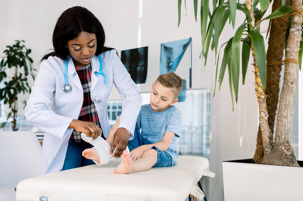 Putting bandage. Dark-skinned woman pediatrician wearing white coat putting bandage on leg of little boy - Фото, изображение