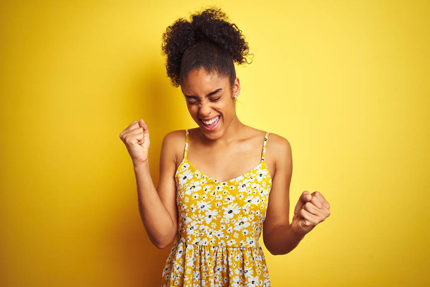 African american woman wearing casual floral dress standing over isolated yellow background very happy and excited doing winner gesture with arms raised, smiling and screaming for success. Celebration concept. - 写真・画像