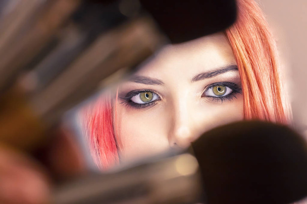 Молодая красивая девушка с рыжими волосами смотрит через щетки для макияжа. Яркие глаза крупным планом. Концепция косметических услуг, уход за кожей, современная мода, макияж, новая звезда знаменитости
 - Фото, изображение