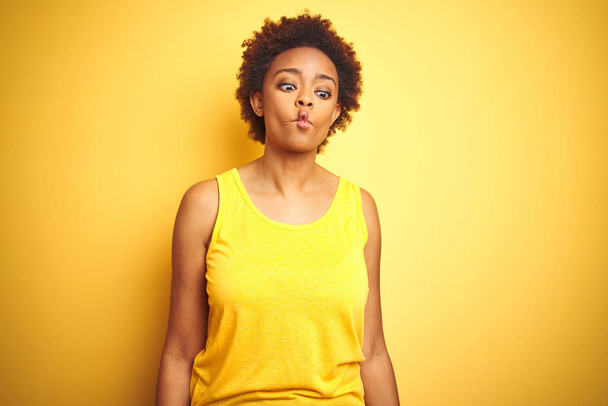 孤立した黄色の背景に夏のTシャツを着た美しいアフリカ系アメリカ人女性は、唇、狂気とコミカルなジェスチャーで魚の顔を作ります。面白い表現. - 写真・画像