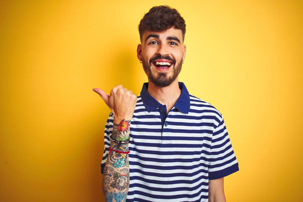 Nuori mies tatuointi yllään raidallinen poolo seisoo yli eristetty keltainen tausta hymyillen onnellinen kasvot etsivät ja osoittaa sivuun peukalo ylöspäin
. - Valokuva, kuva